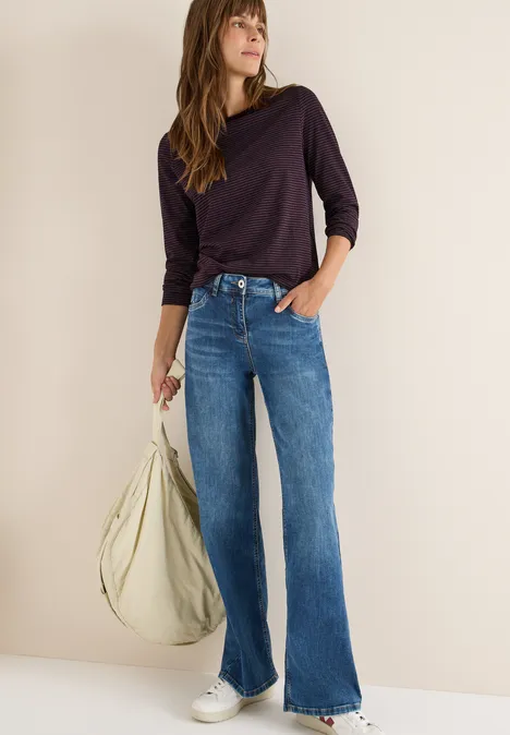 Frau mit gestreiftem Shirt, Jeans und Tasche steht locker an einer Wand.